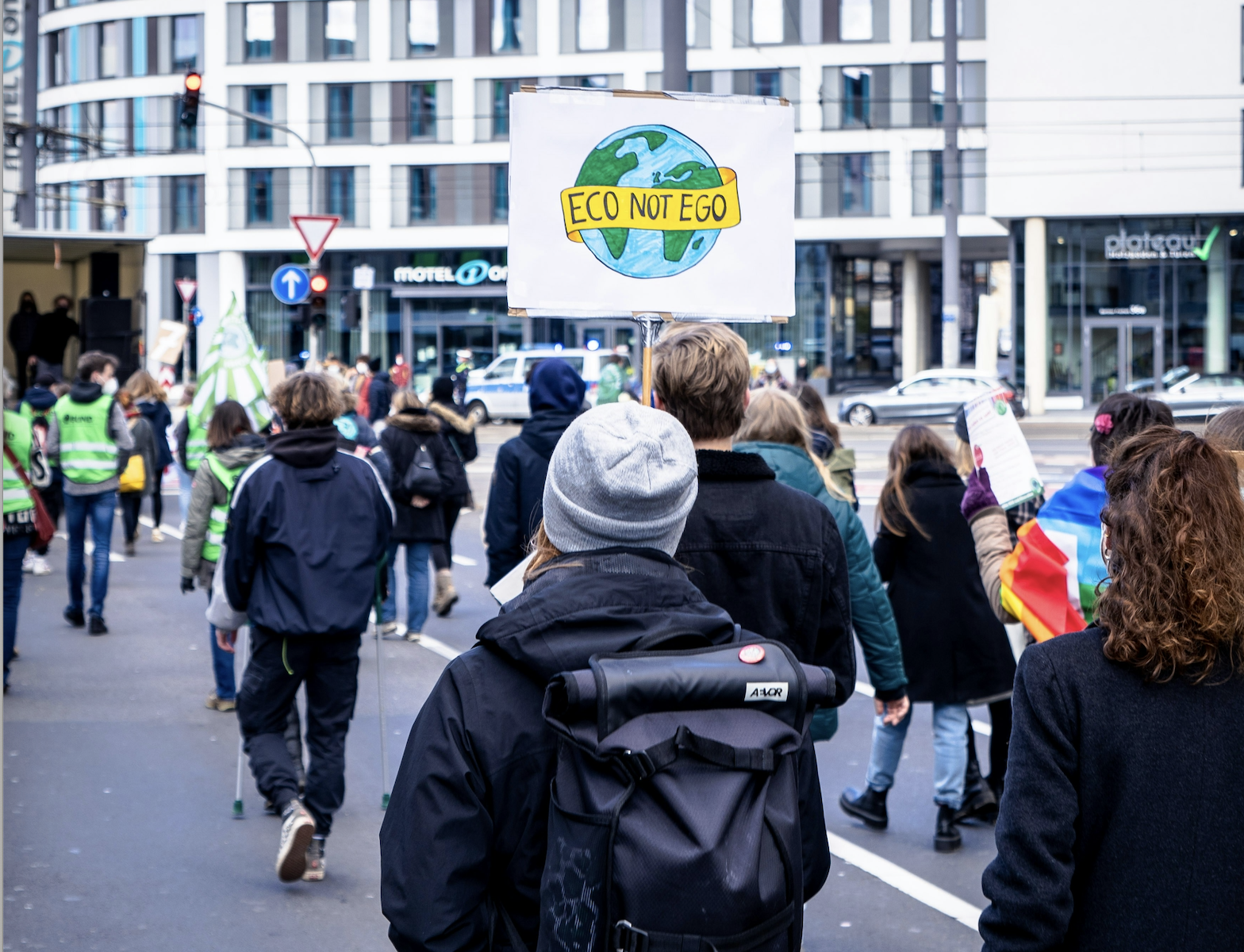 people in winter attire walking at a climate protest, one carrying a sign that reads "Eco Not Ego" written on a yellow banner with the image of a globe behind it